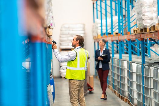 Side view of Caucasian male worker scanning package with barcode scanner in modern warehouse. Diverse staff working behind him in the aisle of the modern warehouse. This is a freight transportation and distribution warehouse. Industrial and industrial workers concept