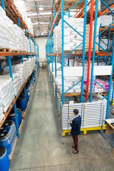 High angle view of mature Caucasian female manager working on white board in warehouse. In the end of the aisle in warehouse diverse staff are working. This is a freight transportation and distribution warehouse. Industrial and industrial workers concept