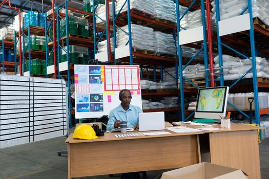 Front view of mature African-american male supervisor working on laptop at desk in warehouse. This is a freight transportation and distribution warehouse. Industrial and industrial workers concept