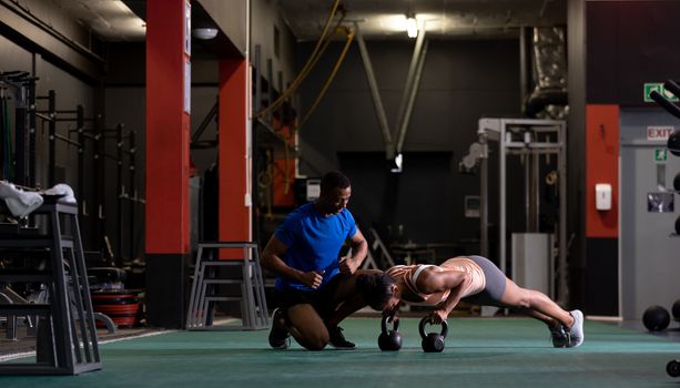 Side view of an African-American woman doing push ups with ball weights and an African-American man assisting her inside a room at a sports centre. Bright modern gym with fit healthy people working out and training. Bright modern gym with fit healthy people working out and training