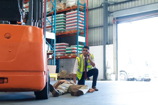 Front view of Caucasian Male supervisor talking on mobile phone while his coworker lying unconscious on the floor in warehouse. This is a freight transportation and distribution warehouse. Industrial and industrial workers concept