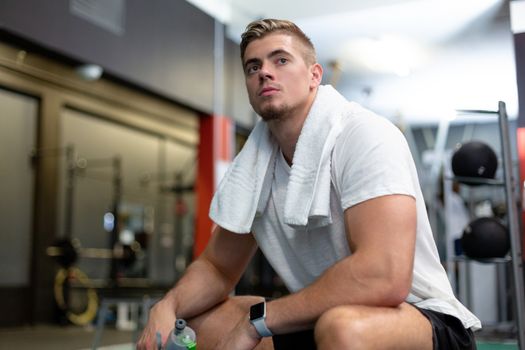 Front view of young thoughtful Caucasian male athletic relaxing in fitness center. Bright modern gym with fit healthy people working out and training