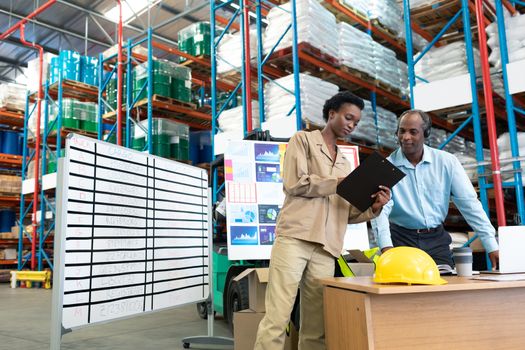Front view of mature African-american male supervisor with beautiful young African-american female coworker discussing over clipboard at desk in warehouse. This is a freight transportation and distribution warehouse. Industrial and industrial workers concept