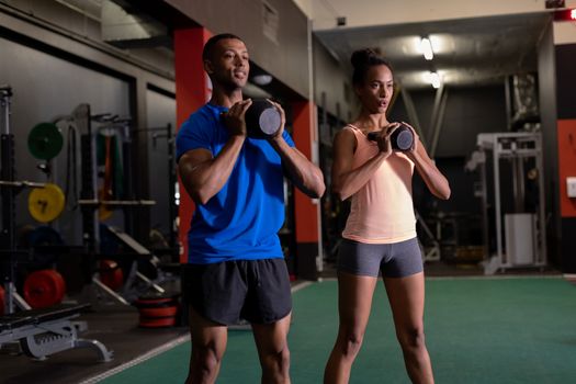 Front view of an African-American man and woman lifting ball weights with both arms inside a room at a sports centre. Bright modern gym with fit healthy people working out and training. Bright modern gym with fit healthy people working out and training