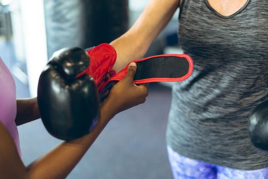 Close-up of young African-american female trainer helping Senior Caucasian female athletic to wear boxing gloves in fitness center. Bright modern gym with fit healthy people working out and training