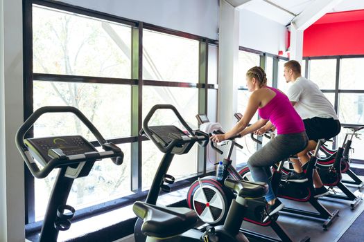 Rear view of young Caucasian male and female athlete exercising with exercise bike in fitness center. Bright modern gym with fit healthy people working out and training