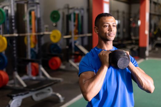 Front view of an African-American man lifting ball weights with both arms inside a room at a sports centre. Bright modern gym with fit healthy people working out and training. Bright modern gym with fit healthy people working out and training