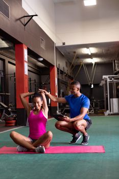 Front view of an African-American woman stretching while sitting crossed legged and an African-American man assists her inside a room at a sports center. Bright modern gym with fit healthy people working out and training. Bright modern gym with fit healthy people working out and training