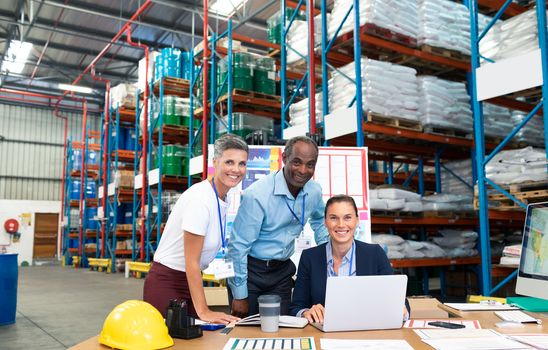 Portrait of beautiful Caucasian female manager with her coworkers discussing over laptop at desk in warehouse. This is a freight transportation and distribution warehouse. Industrial and industrial workers concept