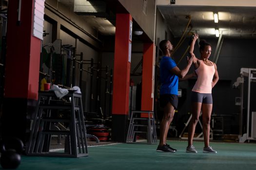 Front view of an African-American woman lifting ball weights and an African-American man assisting her inside a room at a sports centre. Bright modern gym with fit healthy people working out and training. Bright modern gym with fit healthy people working out and training