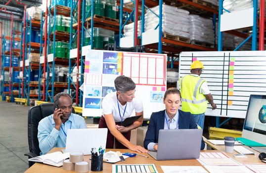 Front view of mature Caucasian female manager with her coworker discussing over laptop at desk in warehouse. This is a freight transportation and distribution warehouse. Industrial and industrial workers concept