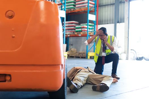 Front view of Caucasian Male supervisor talking on mobile phone while his coworker lying unconscious on the floor in warehouse. This is a freight transportation and distribution warehouse. Industrial and industrial workers concept