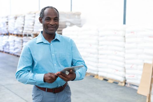 Portrait of happy handsome mature African-american male supervisor looking at camera while working on digital tablet in warehouse. This is a freight transportation and distribution warehouse. Industrial and industrial workers concept