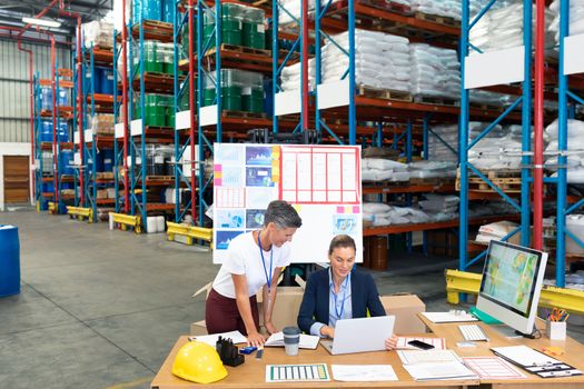 High angle view of beautiful Caucasian female manager with her coworker discussing over laptop at desk in warehouse. This is a freight transportation and distribution warehouse. Industrial and industrial workers concept