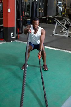 Front view of handsome concentrated fit African-american male athletic exercising with battle ropes in fitness center. Bright modern gym with fit healthy people working out and training