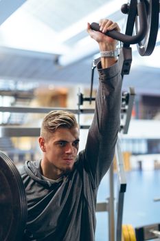 Close-up of handsome young Caucasian man exercising with shoulder machine in fitness studio. Bright modern gym with fit healthy people working out and training