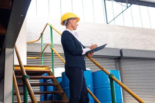 Low angle view of Caucasian female manager writing on clipboard on stairs in warehouse. This is a freight transportation and distribution warehouse. Industrial and industrial workers concept