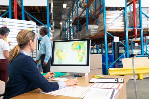 Rear view of pretty mature Caucasian female manager working on computer at desk in warehouse. Diverse colleagues discussing business in front of whiteboard. This is a freight transportation and distribution warehouse. Industrial and industrial workers concept