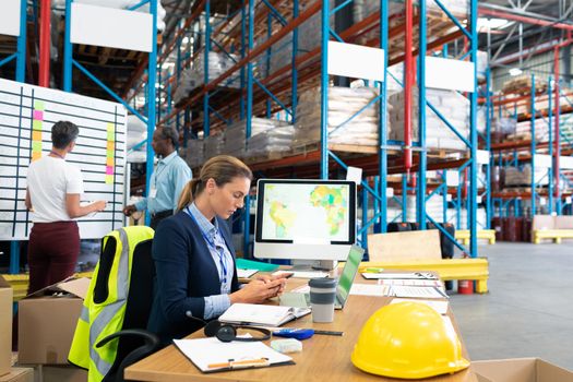 Side view of beautiful Caucasian female manager using mobile phone at desk in warehouse. In the background diverse colleagues are discussing in front of whiteboard. This is a freight transportation and distribution warehouse. Industrial and industrial workers concept