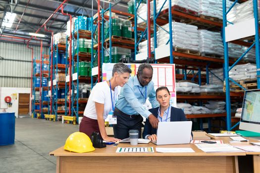 Front view of beautiful Caucasian female manager with her coworkers discussing over laptop at desk in warehouse. This is a freight transportation and distribution warehouse. Industrial and industrial workers concept