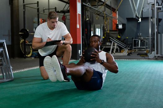 Front view of handsome young Caucasian male trainer writing on clipboard while young fit African-american male athletic exercising with exercise ball in fitness center. Bright modern gym with fit healthy people working out and training