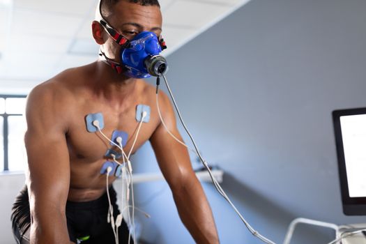 Front view of a naked African-American athletic man doing a fitness test using a mask connected to a monitor while riding an exercise bike inside a room at a sports centre. Bright modern gym with fit healthy people working out and training. Athlete testing themselves with cardiovascular fitness test on exercise bike
