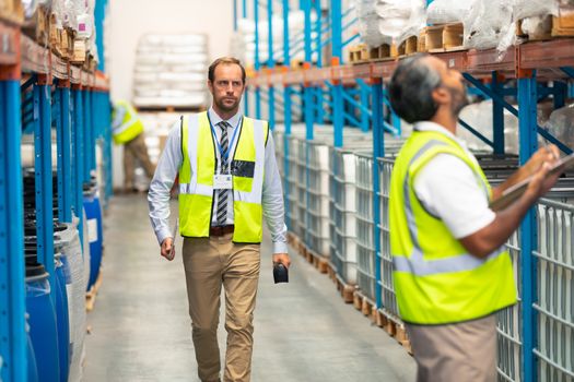 Front view of handsome Caucasian male supervisor walking in warehouse. In front of him an Asian male staff member is writing on clipboard. This is a freight transportation and distribution warehouse. Industrial and industrial workers concept