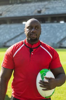 Front view of African American rugby player standing with rugby ball and looking away to the left in stadium on a sunny day.