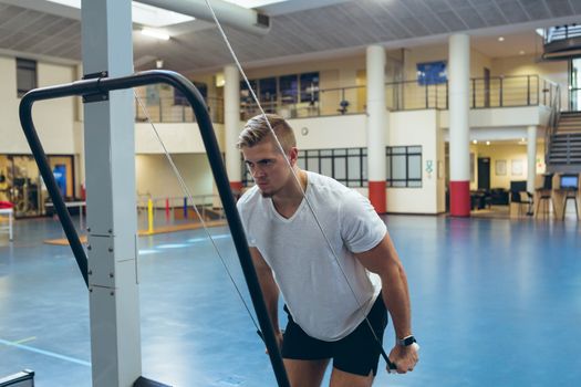 Front view of handsome young Caucasian male athlete doing triceps exercise in fitness studio. Bright modern gym with fit healthy people working out and training