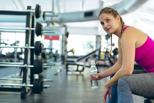 Side view of young fit Caucasian female athletic having water while relaxing on a bench in fitness center. Bright modern gym with fit healthy people working out and training