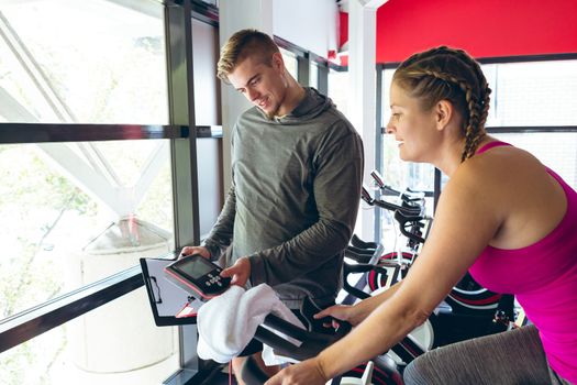 Front view of young Caucasian Male trainer assisting young Caucasian female athlete with exercise in fitness studio. Bright modern gym with fit healthy people working out and training