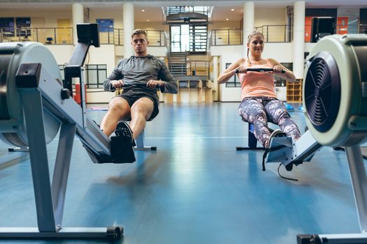 Front view of young Caucasian male and female athlete exercising with rowing machine in fitness studio. Bright modern gym with fit healthy people working out and training