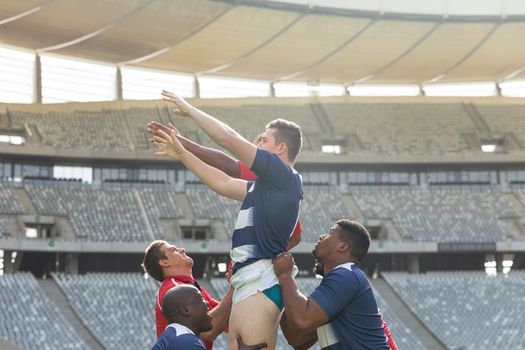 Side view of diverse male rugby players trying to catch the ball in the air while team members hold them in the air in stadium on sunny day.