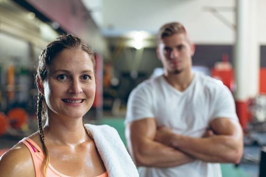 Portrait close-up of pretty Caucasian female athlete looking at camera in fitness center. Handsome young Caucasian man is standing in the background. Bright modern gym with fit healthy people working out and training