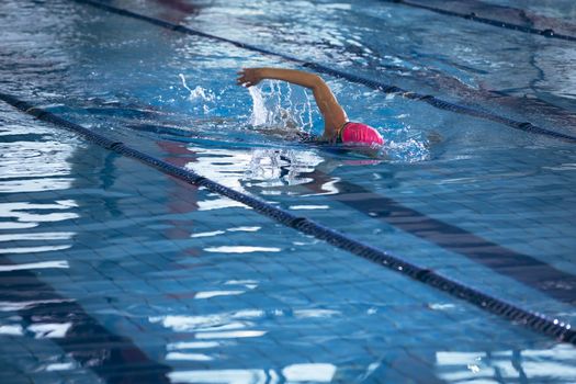 Side view of an African-American woman wearing a pink swimming cap and goggles doing a freestyle stroke in a swimming pool. Bright modern gym with fit healthy people working out and training