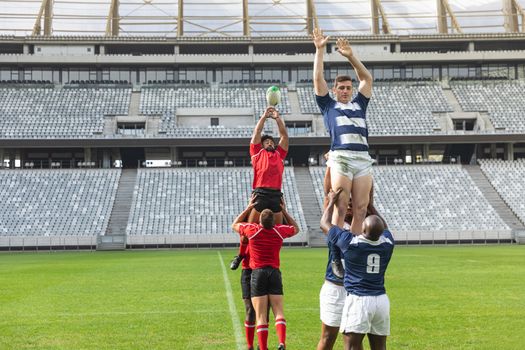 Front view of group of diverse male rugby players playing rugby match in stadium. African American player and Caucasian player are trying to catch the ball in the air.