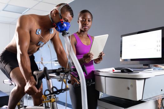 Side view of a naked African-American athletic man doing a fitness test using a mask connected to a monitor while riding an exercise bike and an African-American woman showing him the results inside a room at a sports centre. Bright modern gym with fit healthy people working out and training. Athlete testing themselves with cardiovascular fitness test on exercise bike