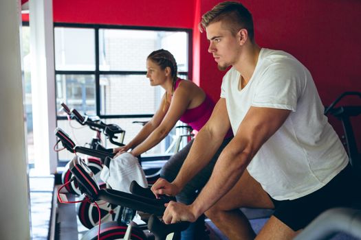 Side view of young Caucasian male and female athlete exercising with exercise bike in fitness center. Bright modern gym with fit healthy people working out and training