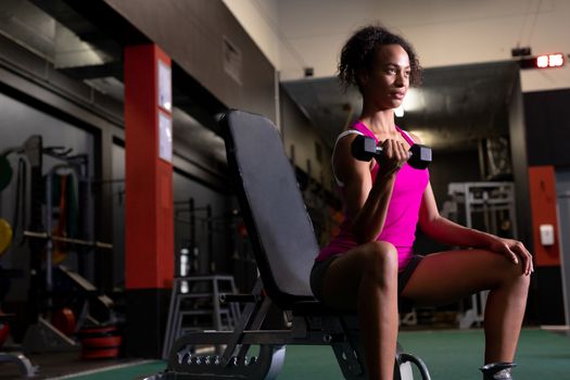 Side view of an African-American woman holding a dumbbell while sitting inside a room at a sports centre. Bright modern gym with fit healthy people working out and training. Bright modern gym with fit healthy people working out and training