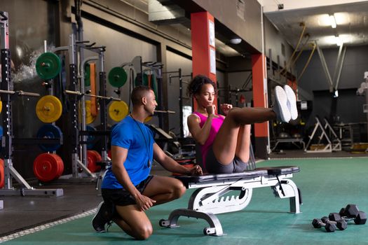 Side view of an African-American woman doing curl ups on a bench and an African-American man assisting her inside a room at a sports center. Bright modern gym with fit healthy people working out and training. Bright modern gym with fit healthy people working out and training