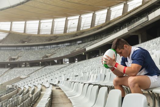 Side view of upset Caucasian male rugby player sitting with rugby ball in stadium