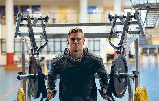 Front view of young fit Caucasian man exercising with shoulder machine in fitness studio. Bright modern gym with fit healthy people working out and training