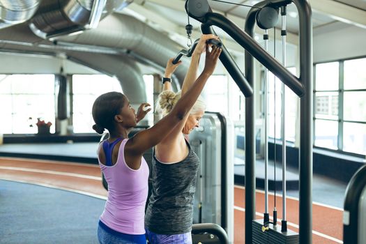 Side view of young pretty African-american female trainer assisting active senior Caucasian  woman on lat pulldown machine in fitness studio. Bright modern gym with fit healthy people working out and training