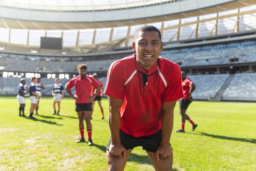 Portrait of tired African American male rugby player standing with hands on his thighs in stadium. Diverse rugby players standing on the rugby field.