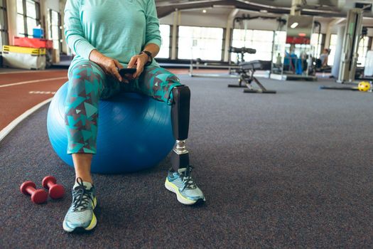 Low section of disabled active senior Caucasian woman with leg amputee using mobile phone while sitting on exercise ball in fitness center. Strong active senior female amputee training and working out