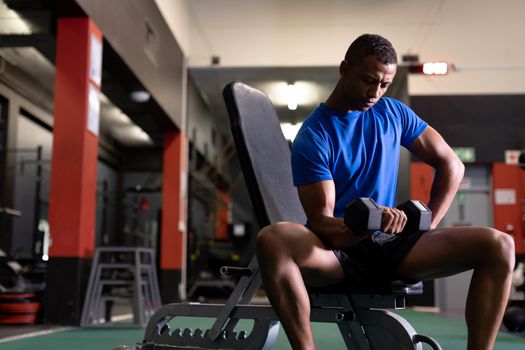 Front view of an African-American man holding a dumbbell while sitting inside a room at a sports centre. Bright modern gym with fit healthy people working out and training. Bright modern gym with fit healthy people working out and training