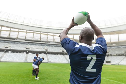 Rear view of African American male rugby player throwing rugby ball in stadium. With players in the background.