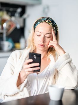 Beautiful caucasian woman at home, feeling comfortable wearing white bathrobe, taking some time to herself, drinking morning coffee and reading news on mobile phone device in the morning.