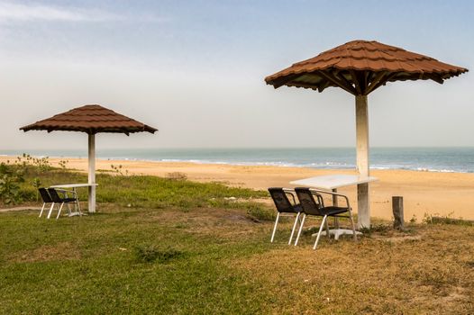 Two chairs and a table are placed facing the sea, just under a red tile umbrella on a beach in Gambia
