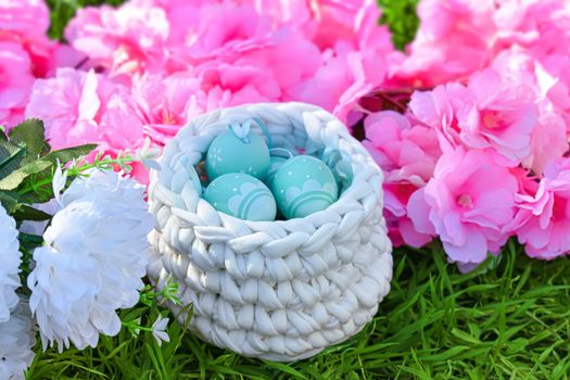 Floral easter background. Painted eggs in crochet basket on green grass. Next to it pink and white flowers decoration.